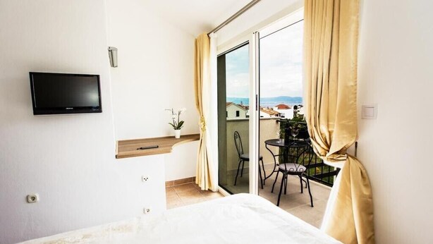 A hotel room with a bed, a TV mounted on the wall, a balcony with a table and chairs, and a view of the cityscape. - Villa Suzy Lux- Villa with Private Pool (Sutivan)