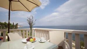 Une table avec un plateau en verre, des chaises blanches et un parasol beige sur un balcon donnant sur la mer.