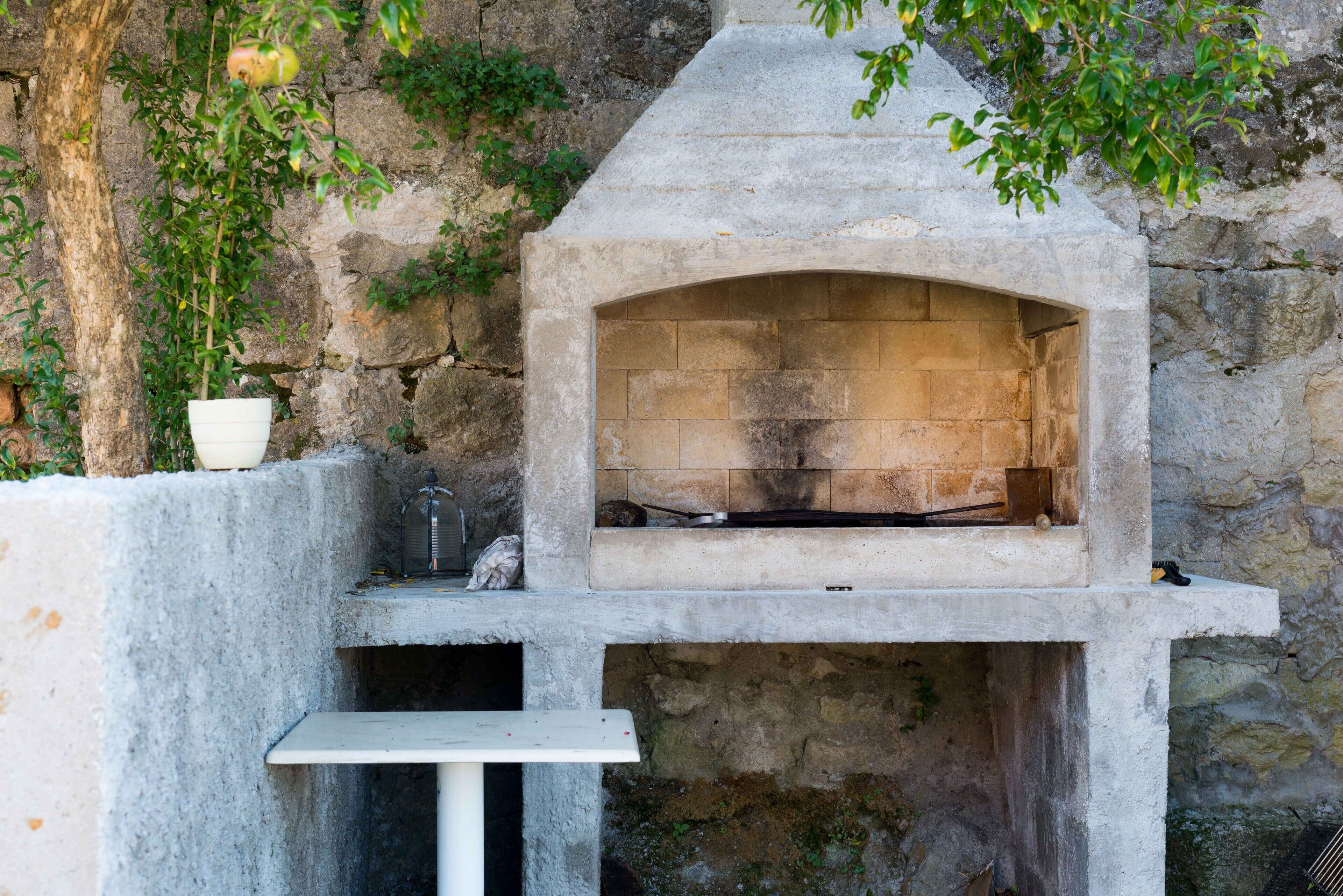 Un horno de piedra al aire libre con un banco en frente, junto a una pared de piedra, un árbol y una planta en maceta.