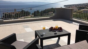 A terrace with wicker furniture, a table with fruit and drinks, and a view of the sea and distant hills. - Apartments Eastern Paradise- Comfort One Bedroom Apartment with Terrace and Sea View (Bol)