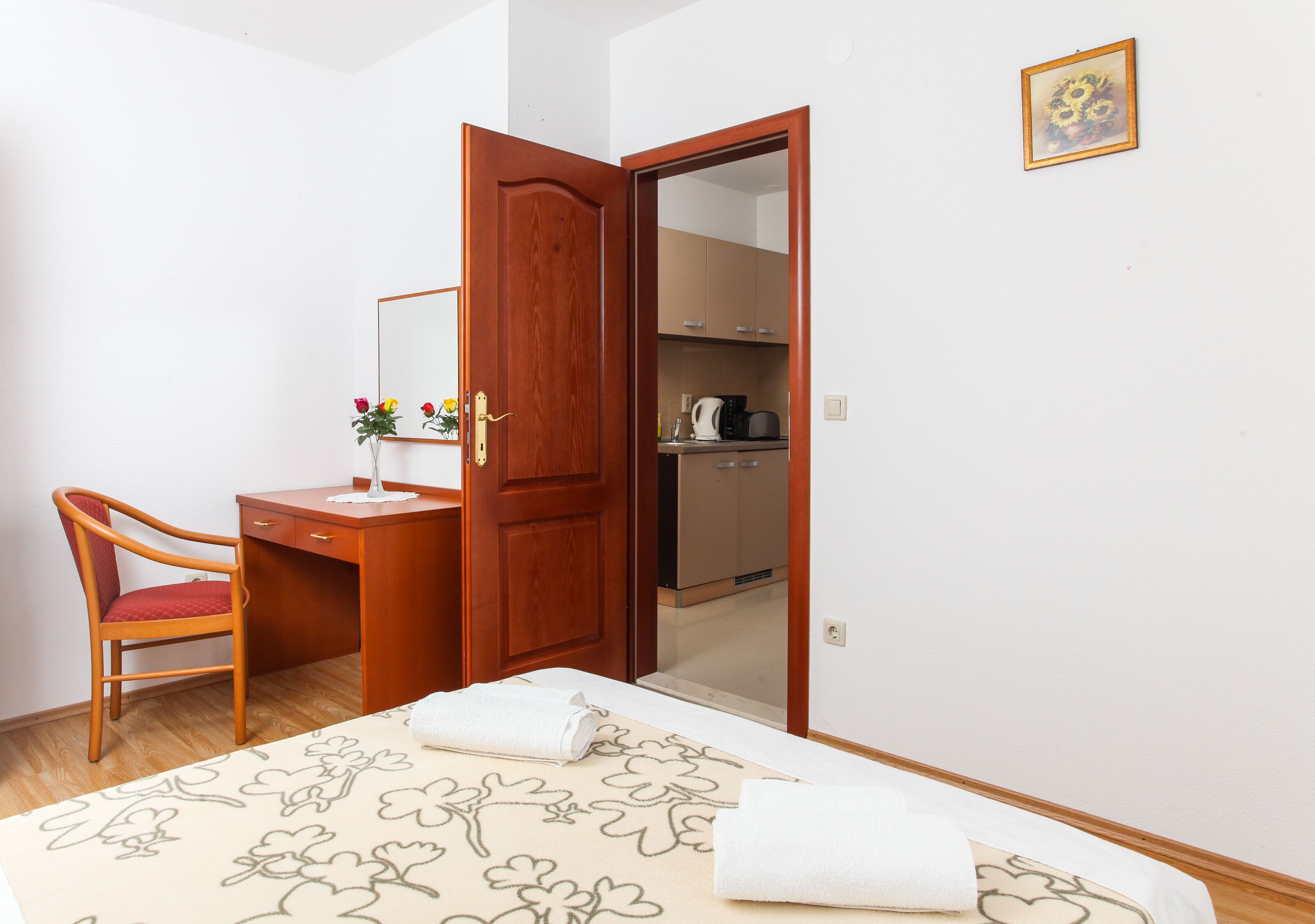 A bedroom with a wooden desk, a chair, a bed with floral bedding, a mirror, and a wardrobe.