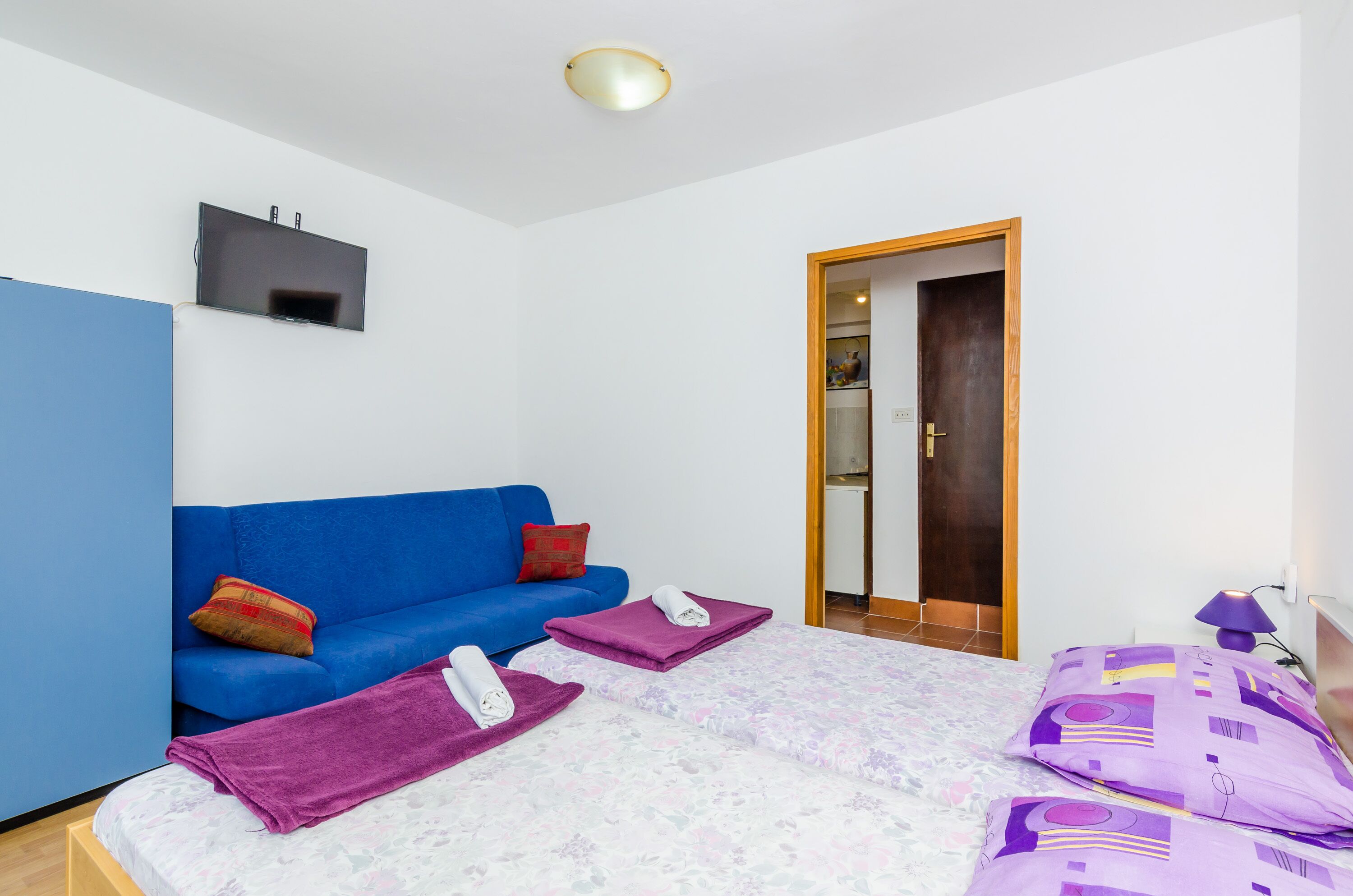 Ein Schlafzimmer mit einem blauen Sofa, einem Bett mit violetter Bettwäsche und einem an der Wand befestigten Fernseher.