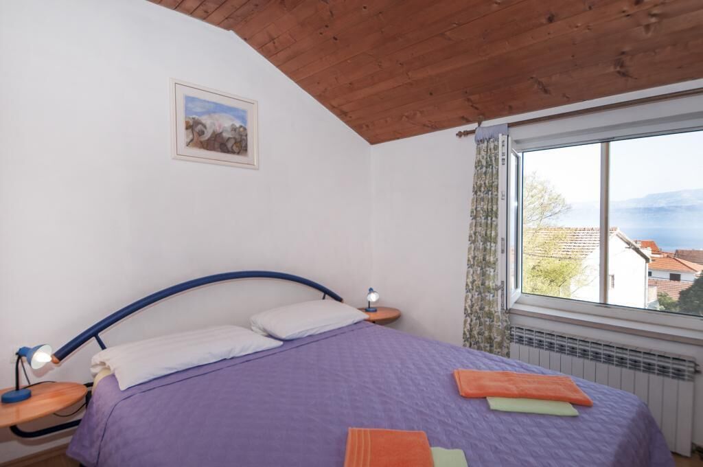 A bedroom with a bed, a nightstand, a lamp, a window with a view, and a painting on the wall.