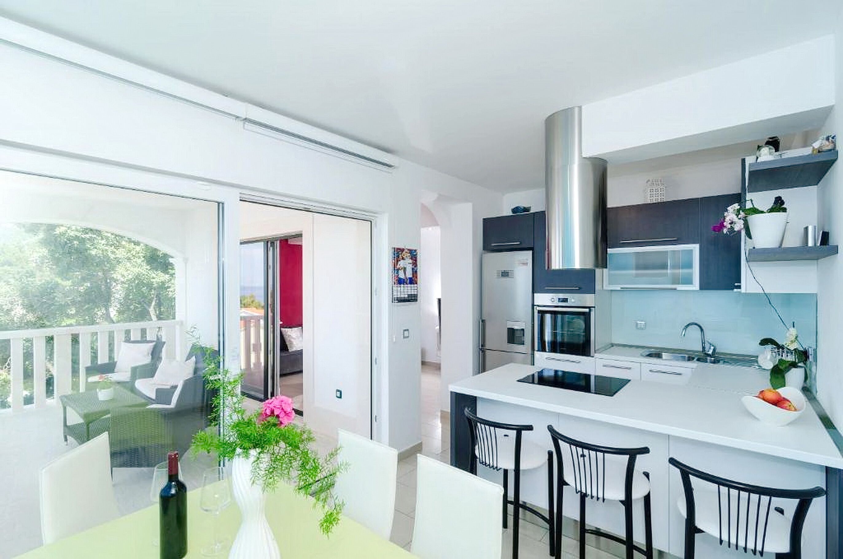 Eine moderne Küche mit weißen Arbeitsflächen, einer Edelstahl-Dunstabzugshaube und einer eingebauten Mikrowelle. Es gibt eine Essecke mit weißem Tisch und Stühlen sowie einen Balkon mit Sitzgelegenheit.