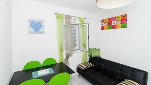 Ein modernes Zimmer mit einer schwarzen Couch, grünen Sesseln, einem schwarzen Tisch, einem Fenster mit grünen Vorhängen, einer weißen Wand mit herzförmiger Dekoration und einem farbenfrohen Kunstwerk.