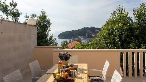 Una mesa dispuesta con frutas, un jarrón de flores y una cafetera en un balcón con vistas a un cuerpo de agua y una ladera.