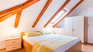 Un dormitorio con techo inclinado, vigas de madera, cama con manta amarilla, mesita de noche con lámpara y armarios blancos.