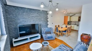 Una sala moderna con una pared de acento de piedra, un televisor de pantalla plana, un sofá, un comedor con mesa y sillas, y una cocina al fondo.
