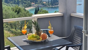 Ein Balkon mit Tisch für zwei Personen, ausgestattet mit einer Obstplatte, zwei Getränken und einer Ananas. Der Ausblick zeigt eine Küstenlandschaft mit Gebäuden und Bäumen.