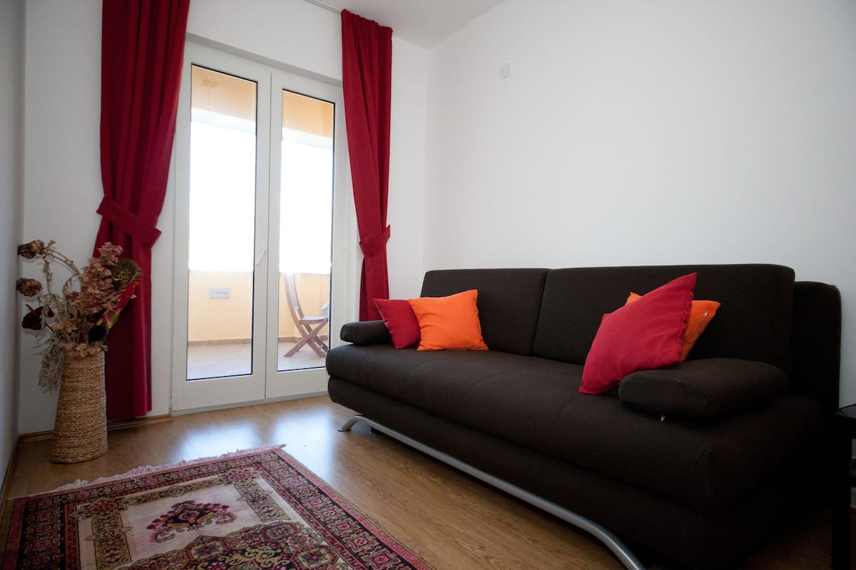 A living room with a dark brown sofa, red and orange cushions, a patterned rug, and a vase with dried flowers.