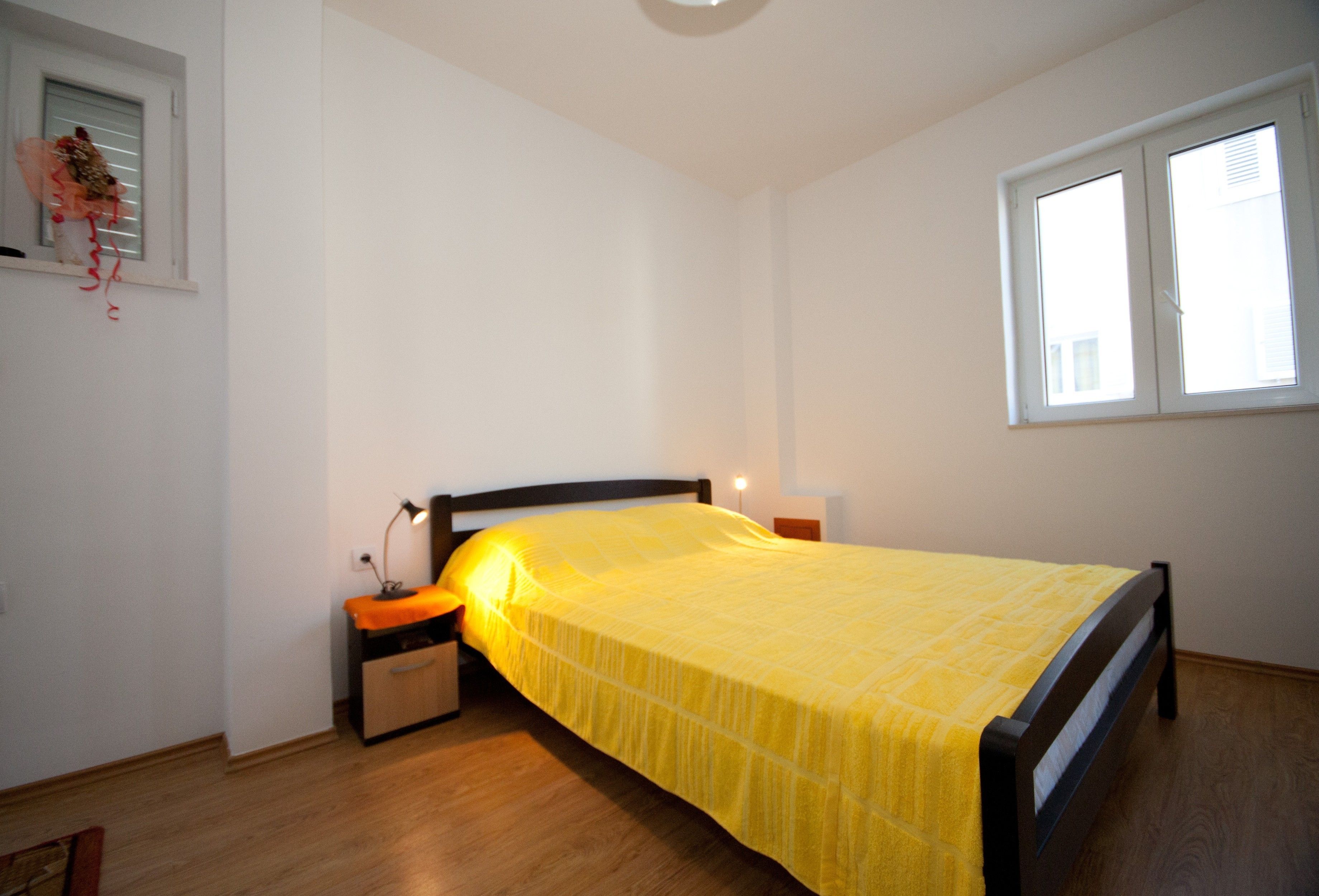 Une chambre avec un plancher en bois, un lit recouvert d’une couvre-lit jaune, une table de chevet avec une lampe et une fenêtre avec des stores.