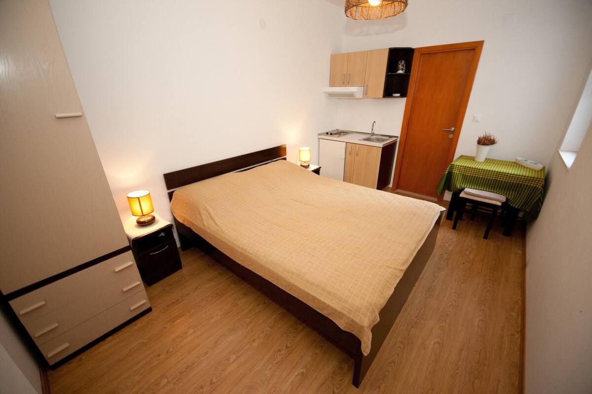 A compact bedroom with a bed, bedside table with a lamp, a small kitchenette, and a wooden door.