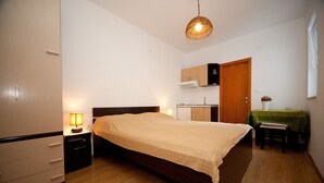A bedroom with a bed, bedside tables, a desk with a chair, a kitchenette, and a door. - Apartments Briest (Dubrovnik)