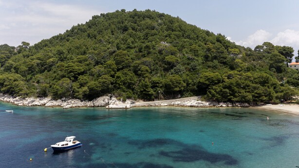 Eine Küstenlandschaft mit einem Boot in einer türkisblauen Bucht, einem Sandstrand und einem bewaldeten Hügel.