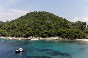 A coastal scene with a boat near a turquoise bay, a sandy beach, and a forested hill.