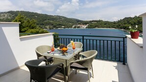 Ein Balkon mit Korbstühlen und einem Tisch, mit Blick auf die Küste, einen klaren Himmel und ferne Hügel.