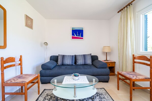 A living room with a blue sofa, two wooden chairs, a glass coffee table with a decorative bowl, a side table with a lamp, and a framed picture on the wall.