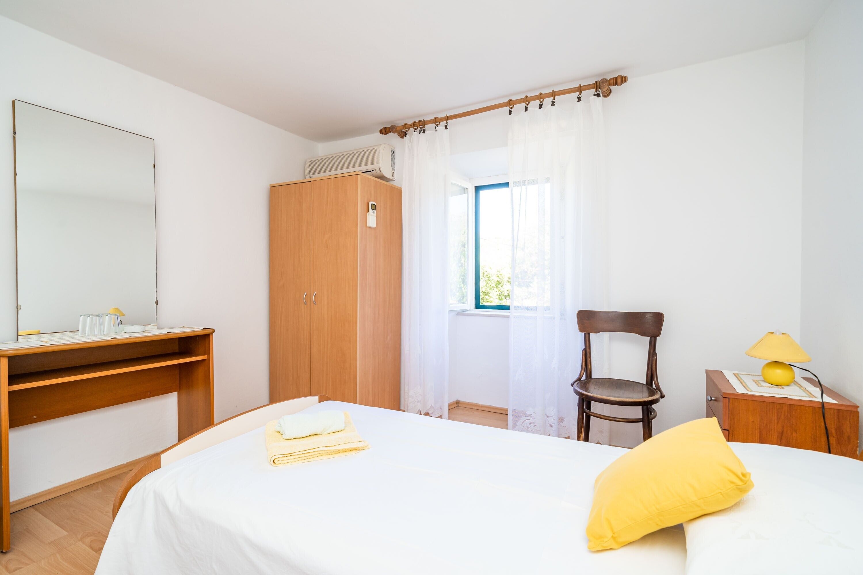 A bedroom with a bed, a wooden wardrobe, a desk with a mirror, a chair, and a bedside table with a lamp.