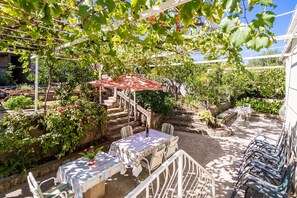A covered outdoor seating area with tables and chairs, surrounded by lush greenery and a pergola.