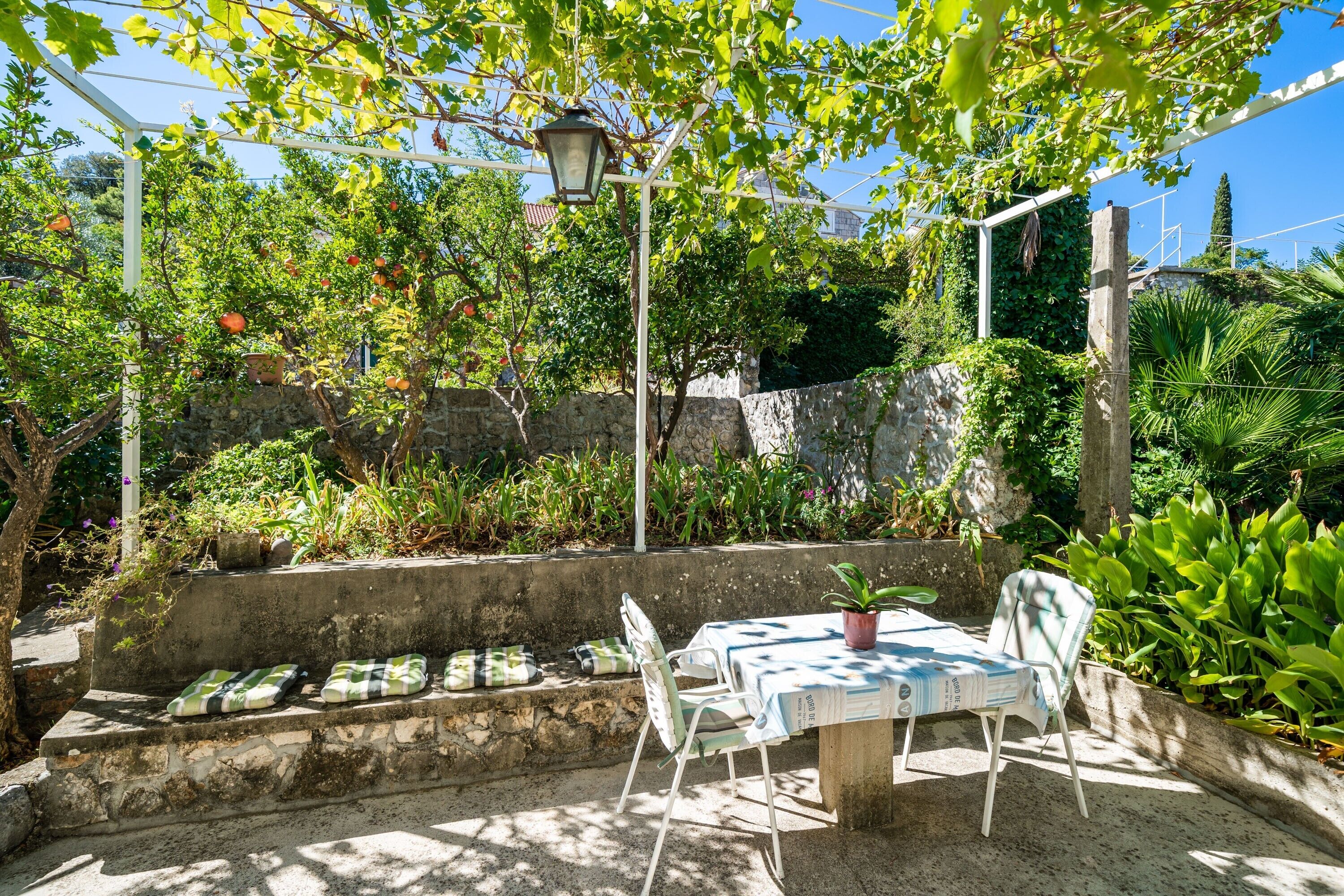 Un patio al aire libre con una mesa y sillas, rodeado de abundante vegetación y un muro de piedra.
