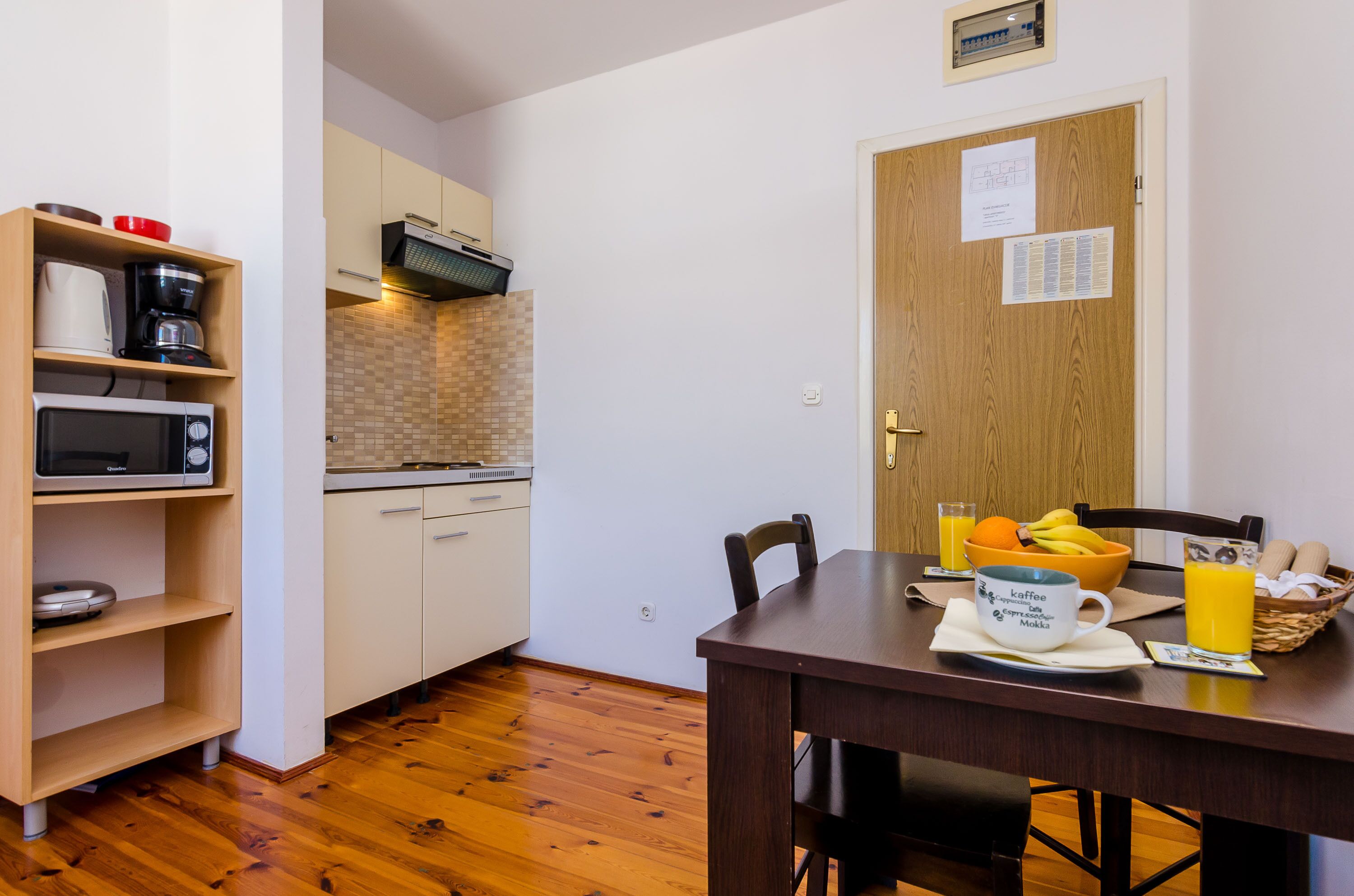 Una pequeña cocina con microondas, cafetera y tostadora en estantes, una mesa de madera con un bol de fruta y una taza de café, y una puerta de madera con un aviso.