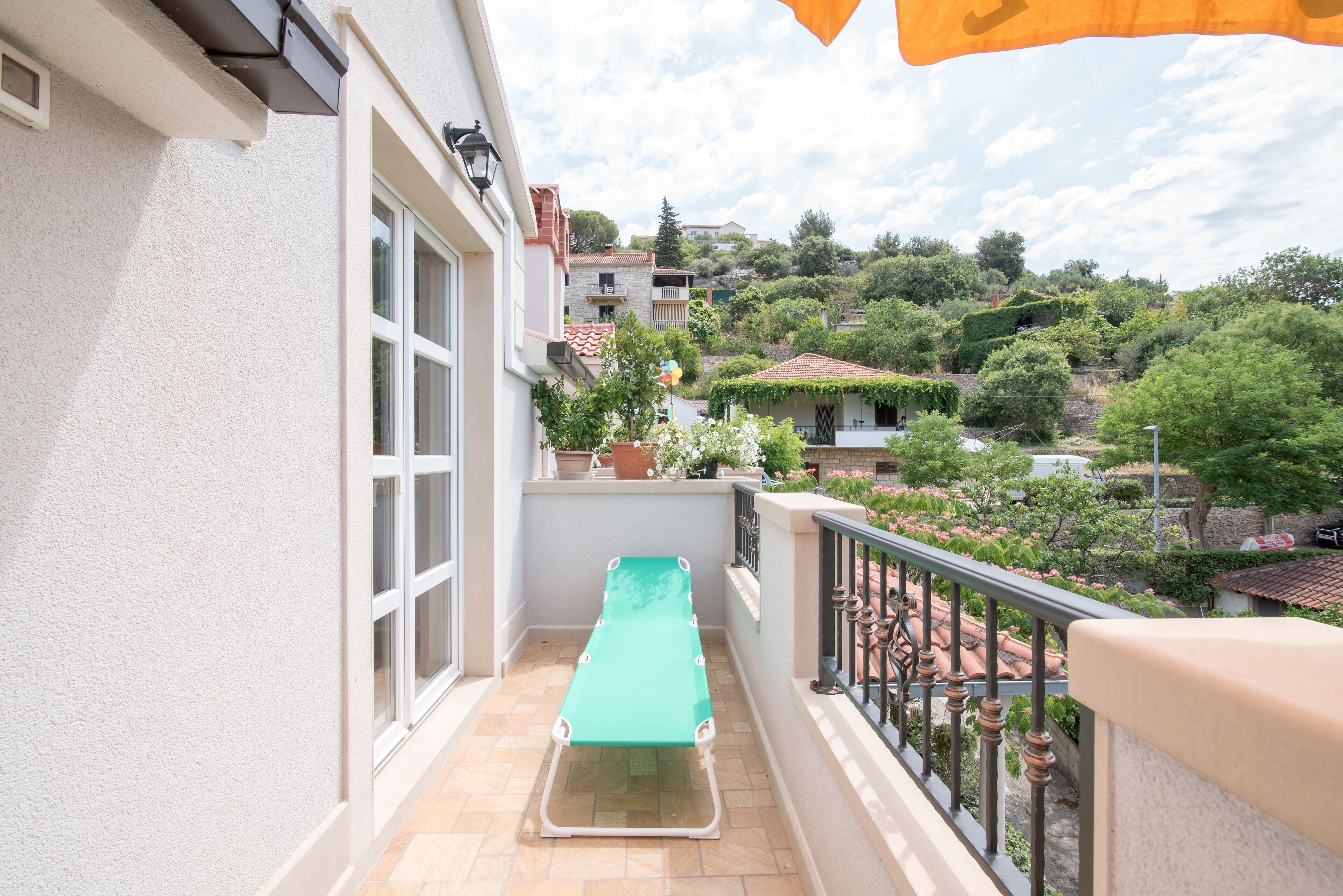 A balcony with a green lounge chair, a potted plant, and a view of a hillside with buildings.