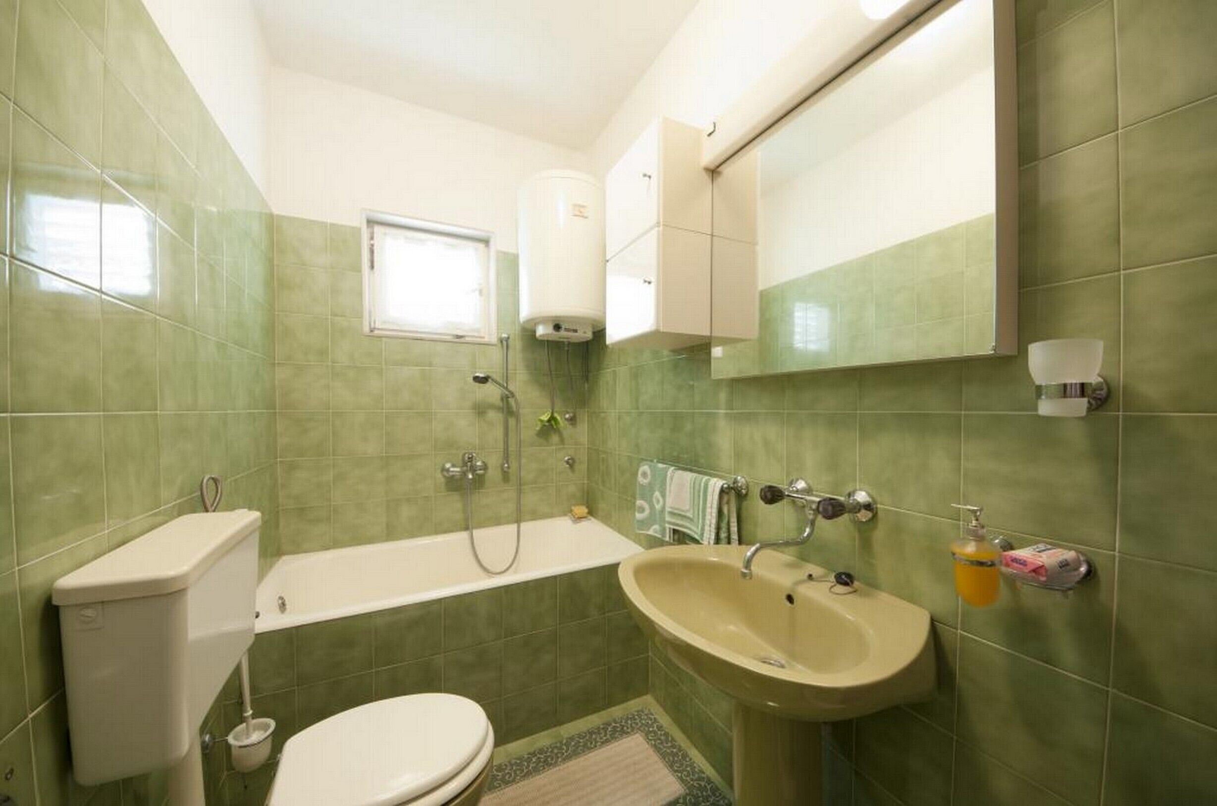 Ein Badezimmer mit grünen Fliesen, einer Toilette, einem Waschbecken, einer Badewanne und einem Fenster.