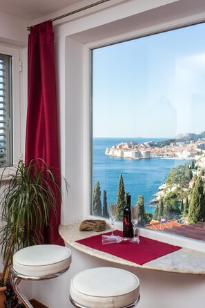 A room with a large window overlooking a coastal city, a red curtain, a plant, two stools, and a bottle of wine on a red mat. - Sea View Apartments - Duplex Four-Bedroom Apartment with Balcony and Sea View - Zlatni Potok 1Street (Dubrovnik)