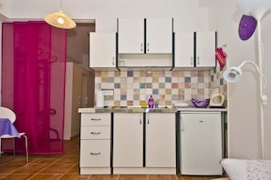 Cozinha compacta com armários brancos, painel de azulejos, geladeira pequena e área de refeições com cadeira e mesa.