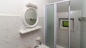 Ein Badezimmer mit einem weißen Waschbecken, einem runden Spiegel, einer Duschkabine aus Glas und einem kleinen Fenster mit Blick auf Grün.