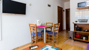 Un espace de vie compact comprenant un téléviseur fixé au mur, un petit coin cuisine avec un micro-ondes et des étagères, ainsi qu’une table à manger avec des chaises.