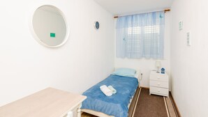 Una habitación pequeña y limpia con una cama individual, una mesita de noche, un tocador y una ventana con cortinas traslúcidas.