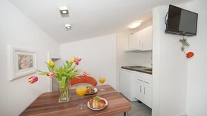 A compact kitchen with white cabinets, a wooden table, a vase with flowers, and a TV mounted on the wall.
