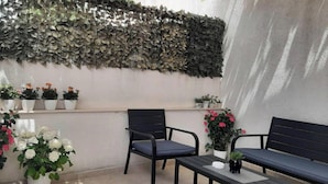 Un patio con una pared cubierta de follaje, varias plantas en macetas y dos sillas negras con una pequeña mesa.