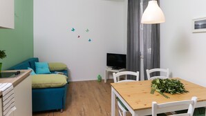 Una stanza piccola e luminosa con un divano, una televisione, un tavolo da pranzo e una finestra con tende.