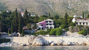 Une propriété située sur la côte, avec une grande maison dotée d’un balcon, entourée d’arbres et bordée d’un rivage rocheux.