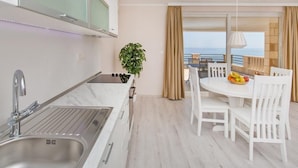 A modern kitchen with a sink, a dining area with white chairs and a table, and a view of the sea through large windows. - Big Blue Apartments - Superior One Bedroom Apartment with Terrace and Sea View (Superior 4) (Dubrovnik)