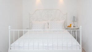 Un lit en métal blanc avec une tête de lit décorative, deux oreillers, une table de chevet avec une lampe et une petite table sur laquelle se trouvent un croissant et un verre de jus d’orange.