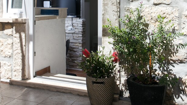 Un bâtiment en pierre avec une porte d’entrée donnant sur une pièce où se trouvent un téléviseur et une petite table. Des plantes en pot sont disposées à l’extérieur de la porte.