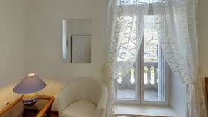 A room with a white chair, a table with a lamp, a window with white curtains, and a view of a building outside.