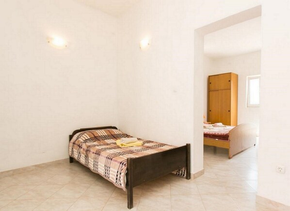 A small, single-bed room with a wooden bed frame and a plaid bedspread. There is a wooden wardrobe in the background and a window providing natural light.