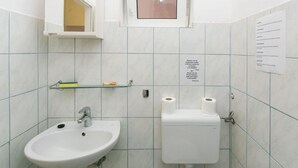 Un baño pequeño con lavamanos, inodoro y un espejo. Hay una ventana sobre el inodoro y un cartel en la pared.