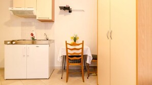 A compact kitchen with white cabinets, a stainless steel countertop, a small table with a chair, and a wall-mounted shelf. - Apartments Villa Ivka - Studio Apartment (Dubrovnik)