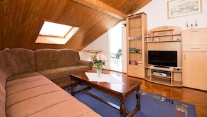 Un accogliente soggiorno con soffitto in legno, una grande finestra, un divano, un tavolino da caffè con un vaso di fiori e una televisione su un mobiletto in legno.