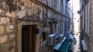 Een smalle, geplaveide steeg met traditionele stenen gebouwen, airconditioners en een houten deur.