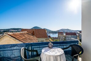 En balkon med et bord og stole, der kigger ud over en by med tage af terracotta og et vandområde.