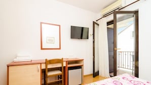 Quarto com cama, escrivaninha, cadeira, frigorífico pequeno, televisão e porta que dá para uma varanda.