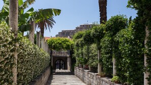 Um caminho de pedra estreito, ladeado por vegetação exuberante e um edifício histórico ao fundo.