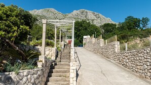 Una escalera de piedra que asciende con una barandilla de metal a un lado y un muro de piedra al otro, rodeada de vegetación y con una montaña al fondo.