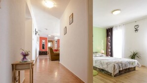 Una habitación de hotel con una cama, una mesita con un florero, una silla y vistas a otra habitación con una pared roja.